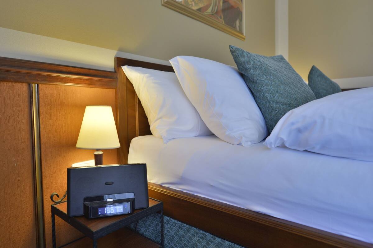 Una cama bien hecha con sábanas blancas y varios almohadones, una mesita de noche con lámpara y un reloj digital, y un cabecero de madera.