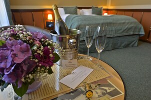 Quarto de hotel com cama, mesa de cabeceira com uma garrafa de champanhe, copos, um buquê de flores e uma placa com o nome.