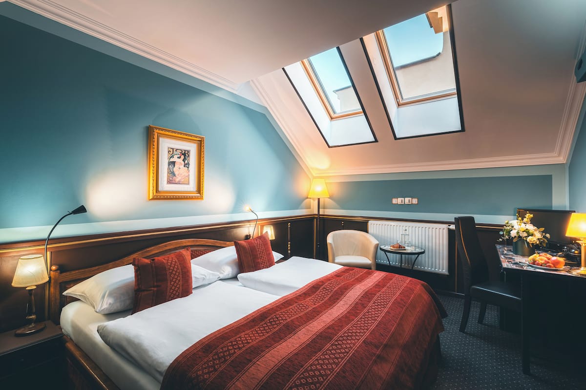 Habitación de hotel con una cama grande, un escritorio y una silla. Hay tragaluces en el techo, un cuadro en la pared y una lámpara sobre el escritorio.
