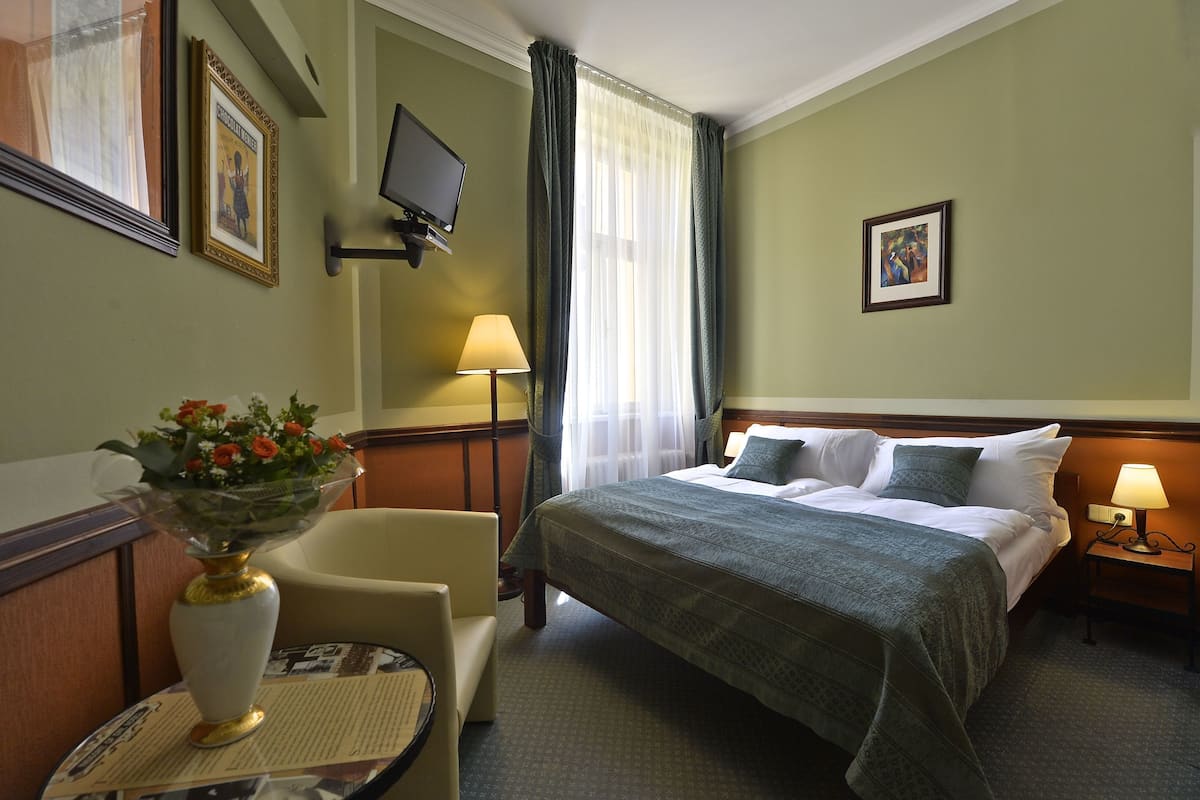 Una habitación de hotel con cama, una silla, una mesa con un jarrón de flores, un televisor montado en la pared y un marco de fotos.