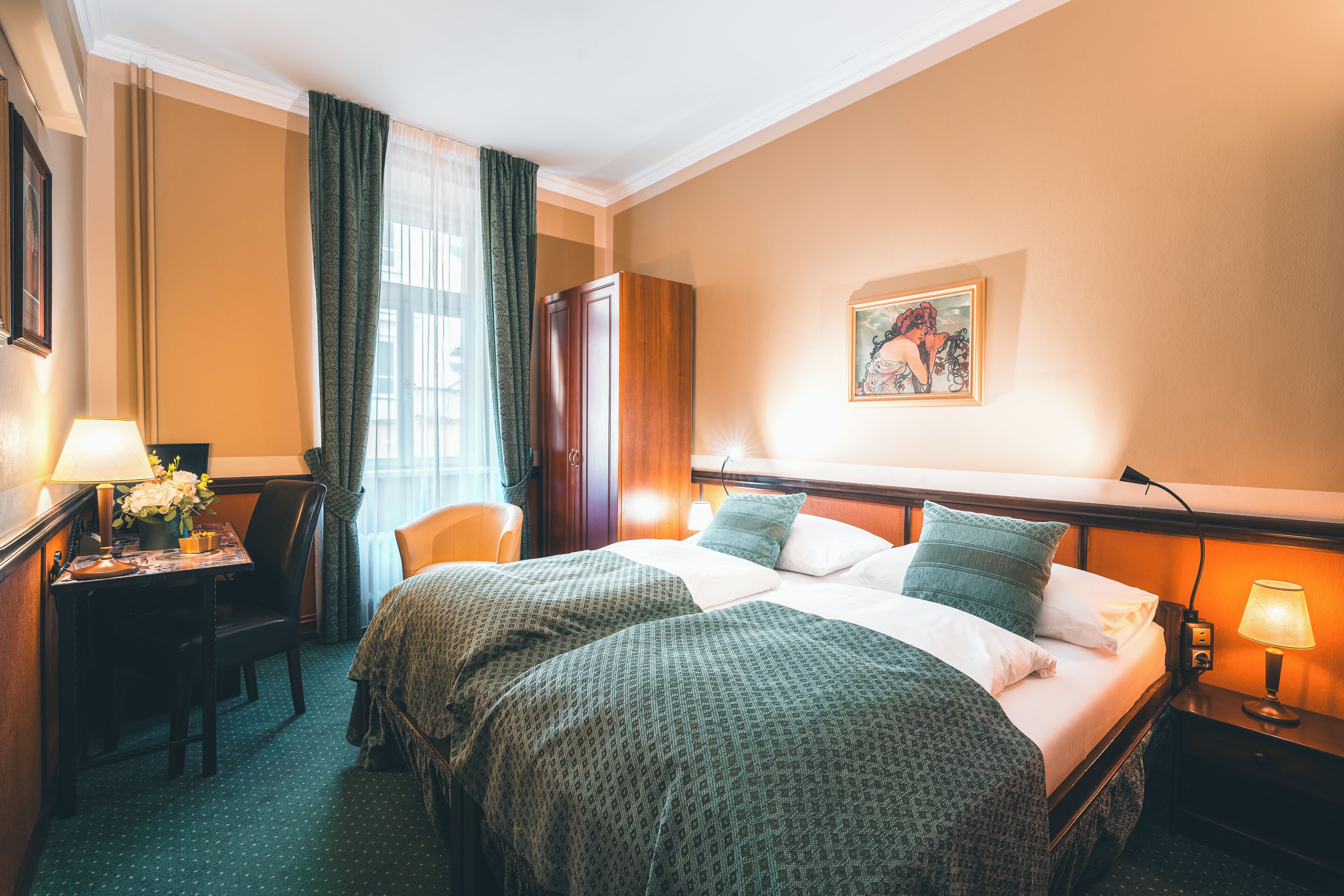 Quarto de hotel com duas camas, uma escrivaninha com abajur, uma cadeira e um quadro na parede.