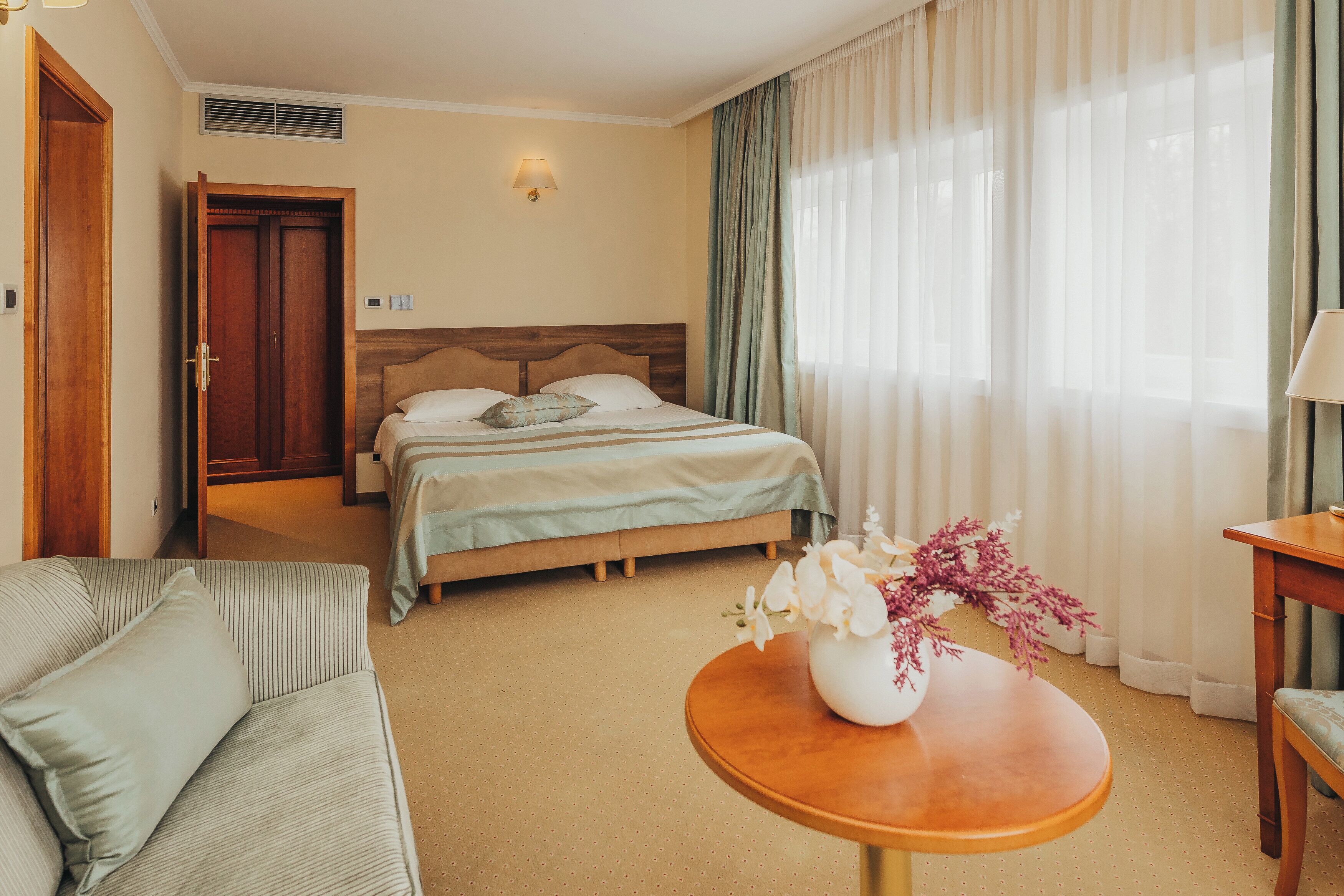A hotel room with a bed, a sofa, a table with a vase of flowers, and a window with curtains.