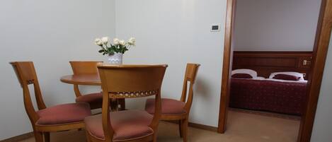 A room with a wooden dining table and four chairs, a vase with white flowers, and a bed with a red bedspread in the background.