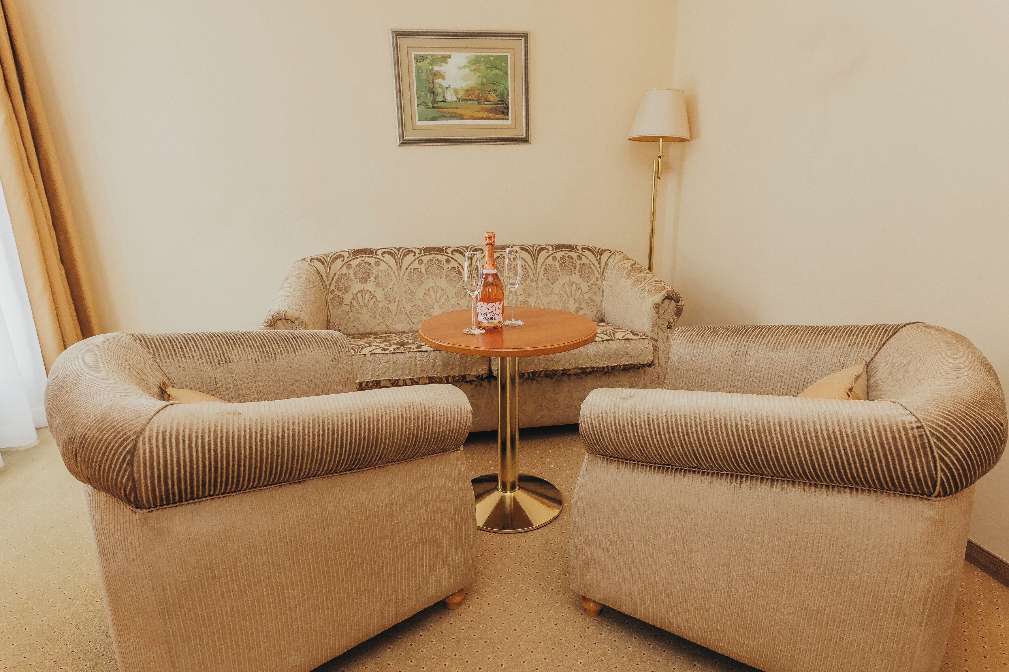 A room with a beige sofa, two armchairs, a small round table, a bottle of wine, and a framed picture on the wall.