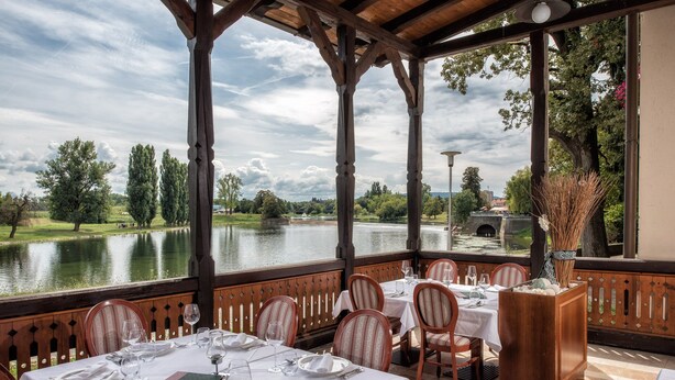 Un espace de restauration en plein air avec des tables disposées pour deux personnes, offrant une vue sur un lac et un parc.