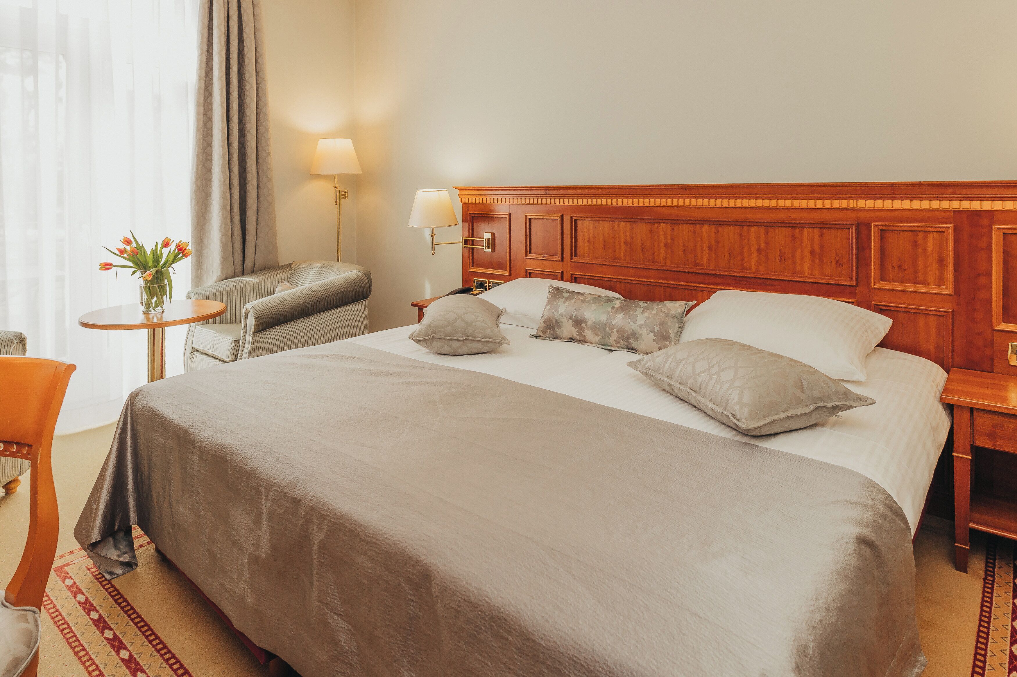 Een hotelkamer met een groot bed, een nachtkastje met een lamp, een stoel en een tafeltje met een bloemenvaas.