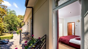 Un balcon avec une table et des chaises, un lit recouvert d’une couverture rouge, et une vue sur un jardin et des voitures garées.