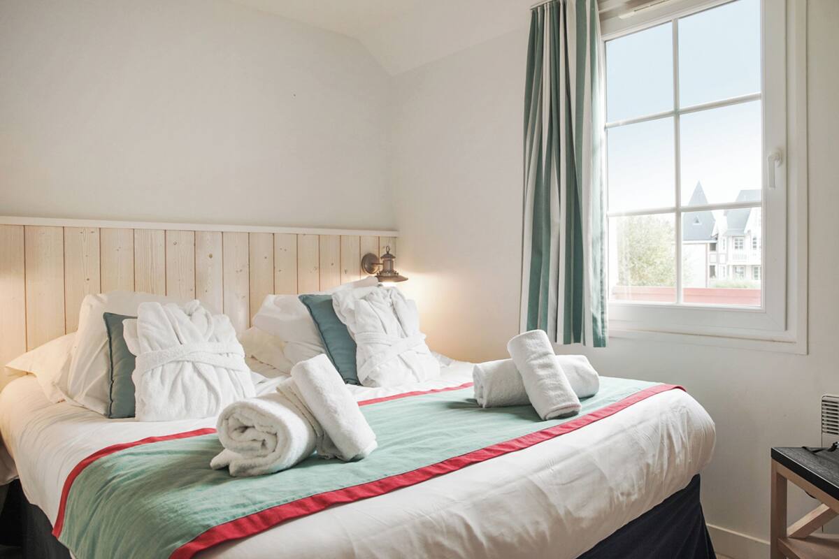 A neatly made bed with white linens, a teal and red striped blanket, and rolled towels.