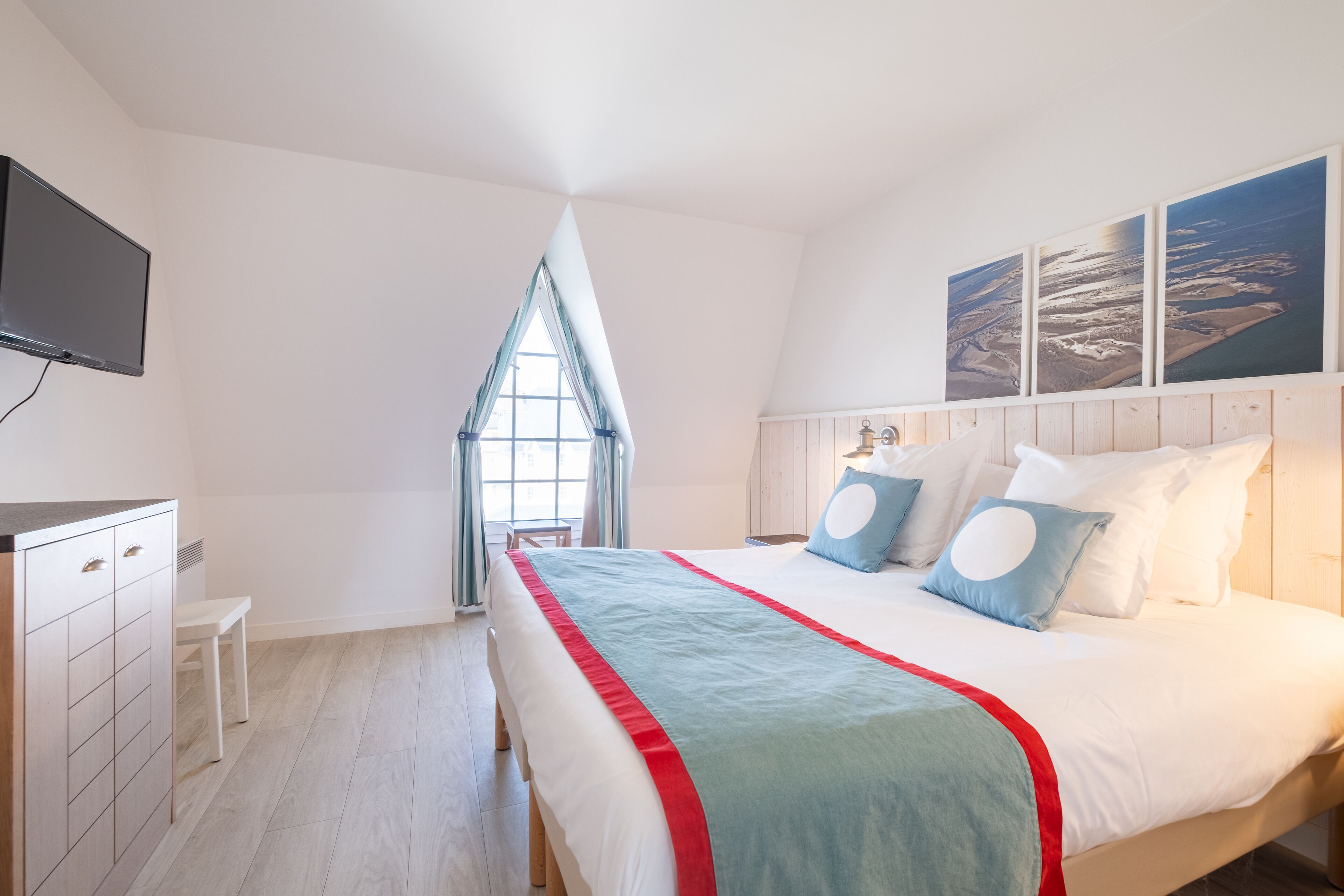 Une chambre à coucher avec un lit, un téléviseur fixé au mur, une fenêtre avec des rideaux et des œuvres d’art au-dessus du lit.