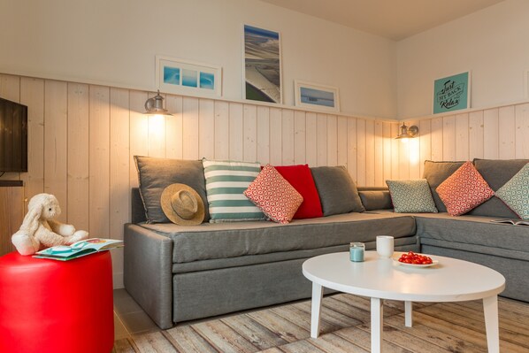 Un salon chaleureux avec un canapé gris, un pouf rouge et une table basse blanche. Des tableaux encadrés ornent le mur et une lampe est posée sur le canapé.