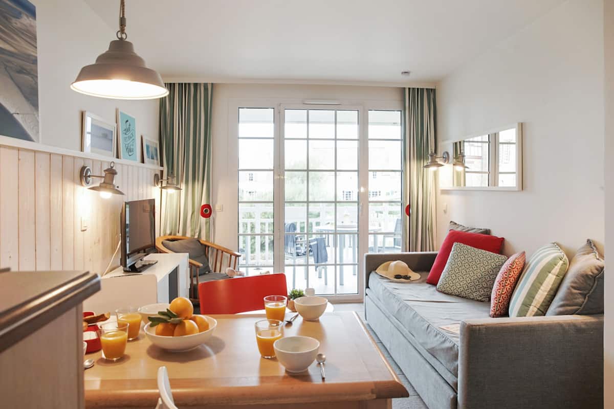 A cozy living room with a sofa, a dining table with breakfast items, and a large window with curtains.