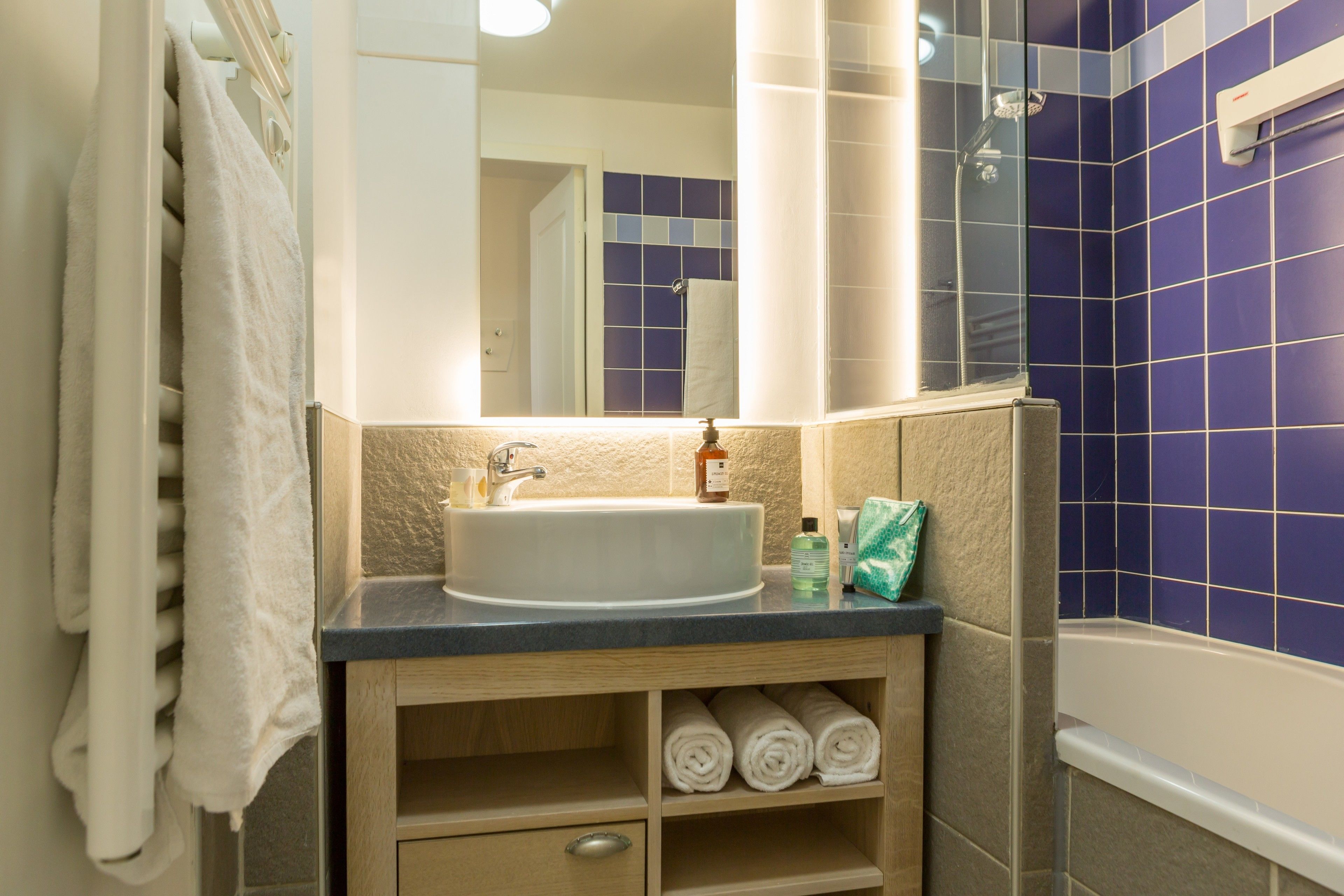 Une salle de bain avec un lavabo, un miroir et des murs recouverts de carreaux bleus. On y trouve des serviettes et des articles de toilette sur le comptoir.