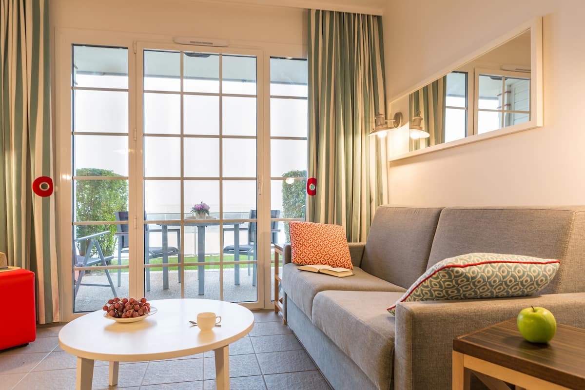 A living room with a sofa, a coffee table with a bowl of grapes and a cup, a wooden side table with an apple, and a sliding glass door leading to an outdoor area.