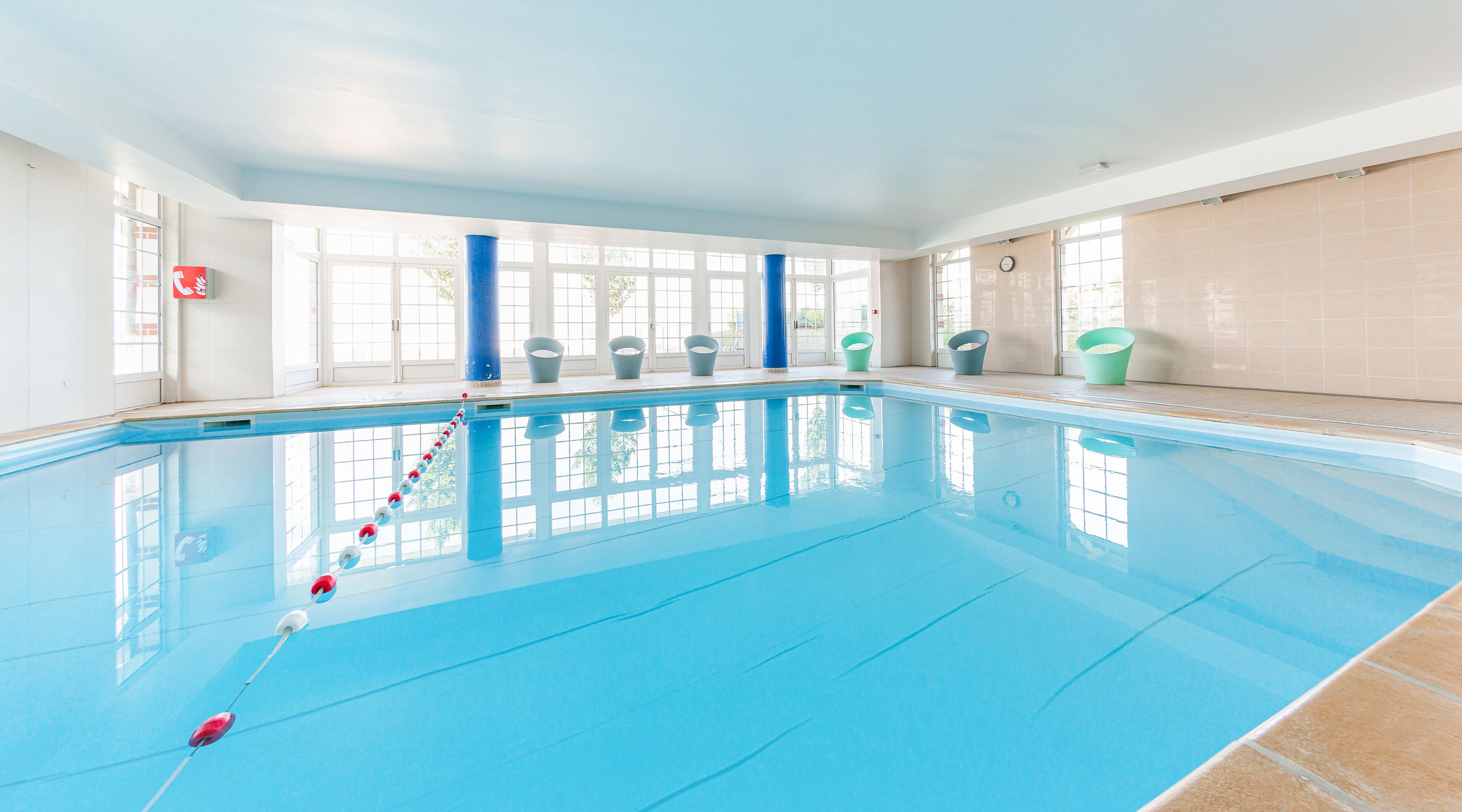 An indoor swimming pool with clear blue water, a starting block, and several empty buckets on the poolside.