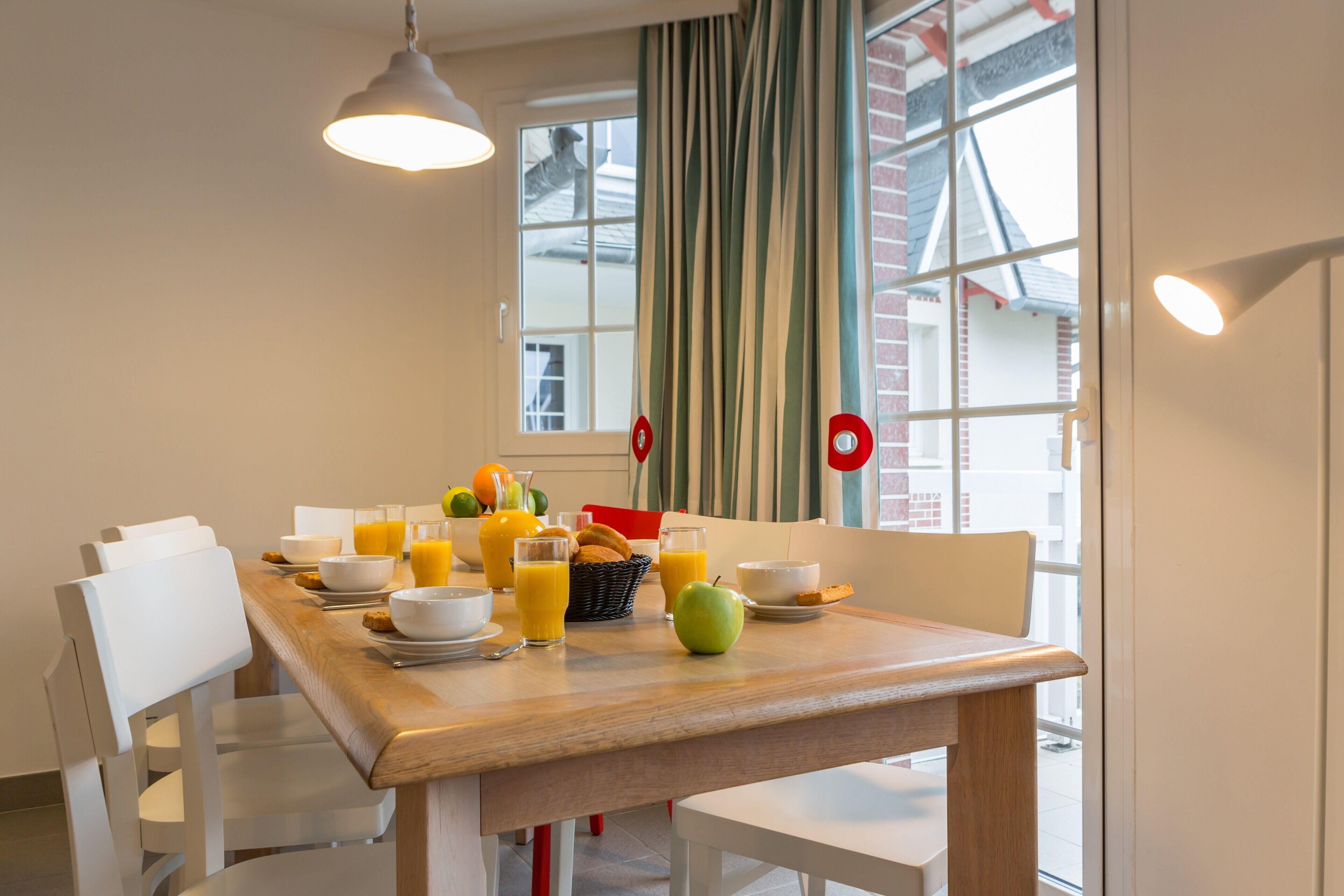 A dining area with a wooden table set for breakfast, white chairs, and a window with curtains.