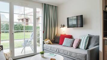 Un salon avec un canapé gris, un téléviseur à écran plat fixé au mur, une porte-fenêtre coulissante donnant sur un jardin, et une petite table ronde sur laquelle se trouvent une plante et un carnet.