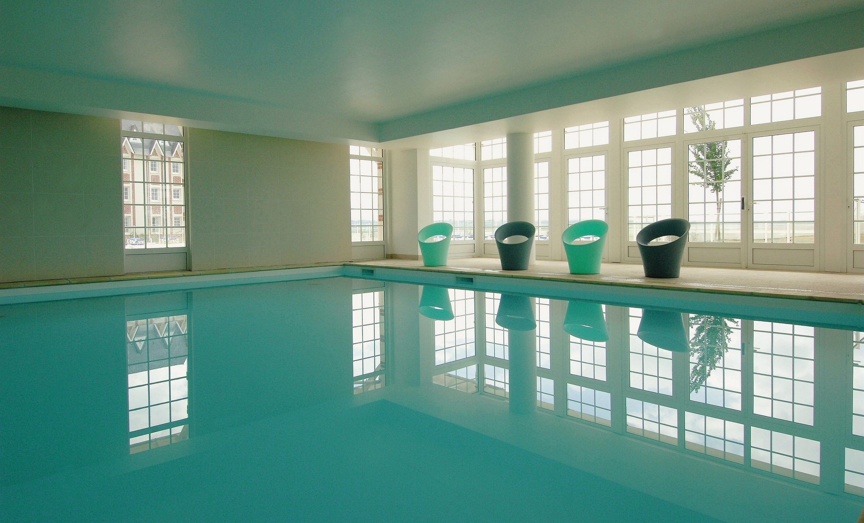 En swimmingpool med klart vatten och en spegelbild av fönstren. Det står fyra stolar på fönsterbrädan.