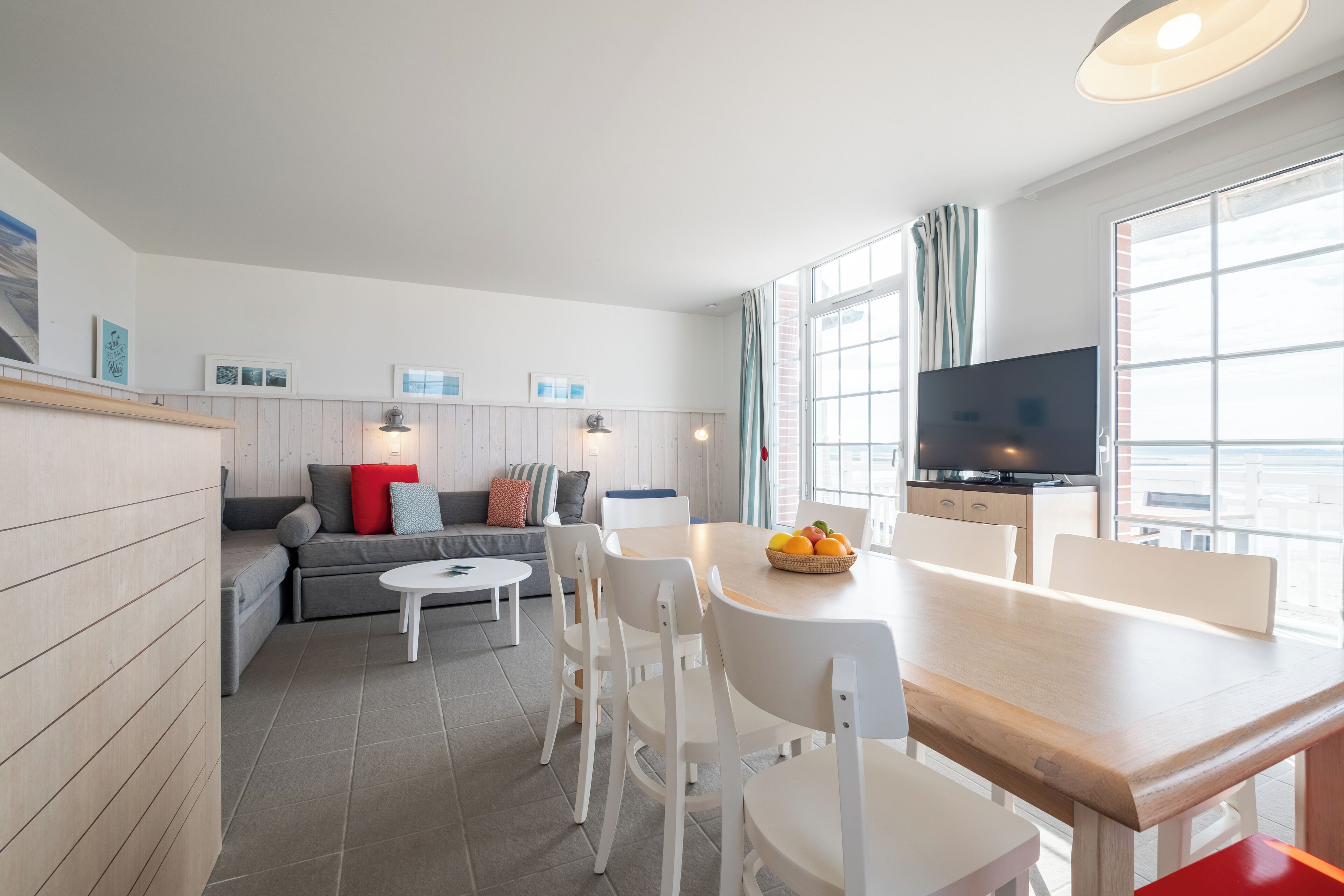 Una sala moderna con sofá, mesa de comedor y sillas. Hay un televisor en la pared y una ventana con cortinas.