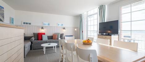 Una sala moderna con sofá, mesa de comedor y sillas. Hay un televisor en la pared y una ventana con cortinas.