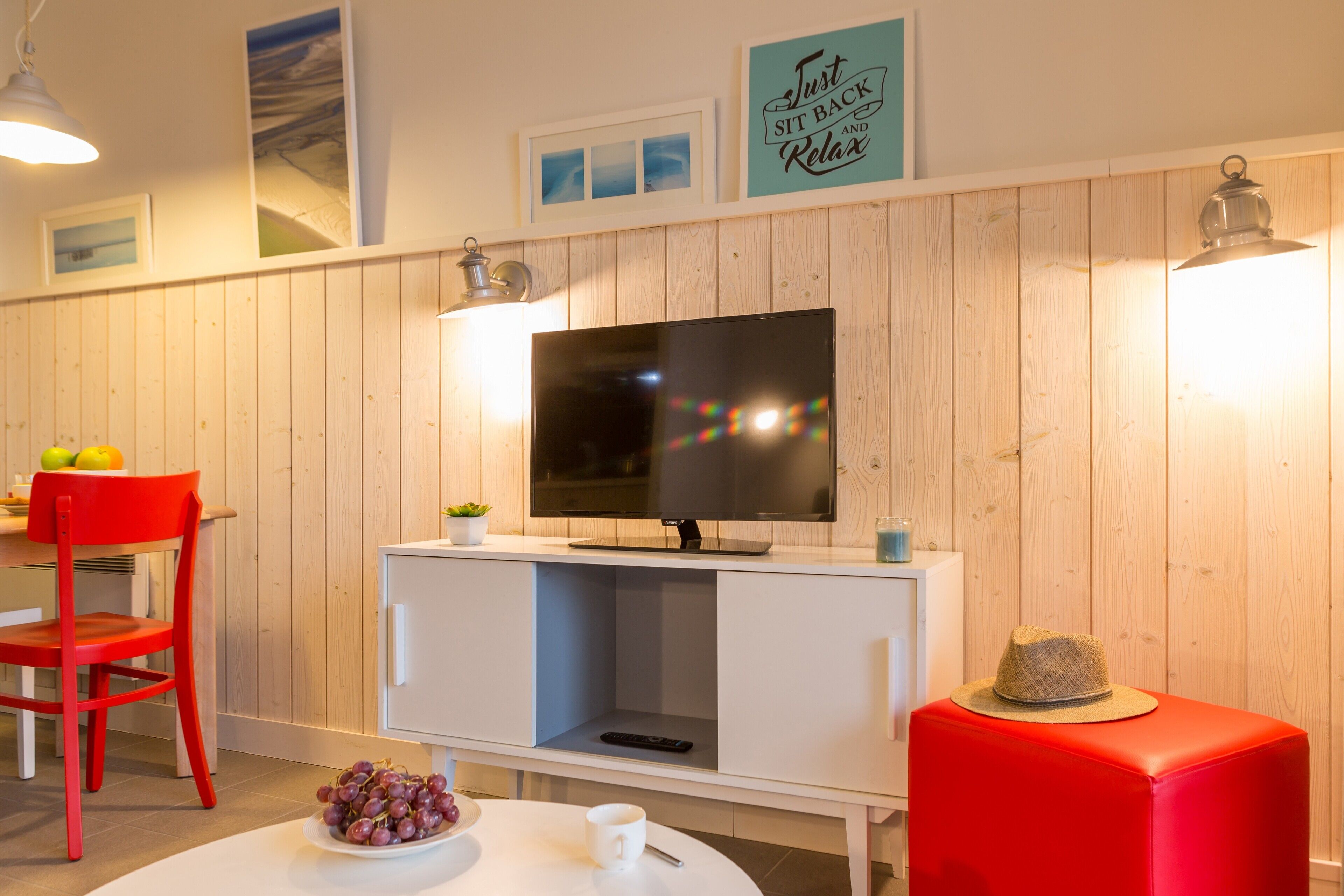 Een kamer met een televisie aan een houten wand, een rode stoel, een witte tafel met een schaal druiven, en een rode poef.