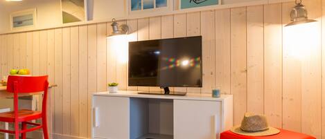 A room with a TV on a wooden wall, a red chair, a white table with a bowl of grapes, and a red ottoman.