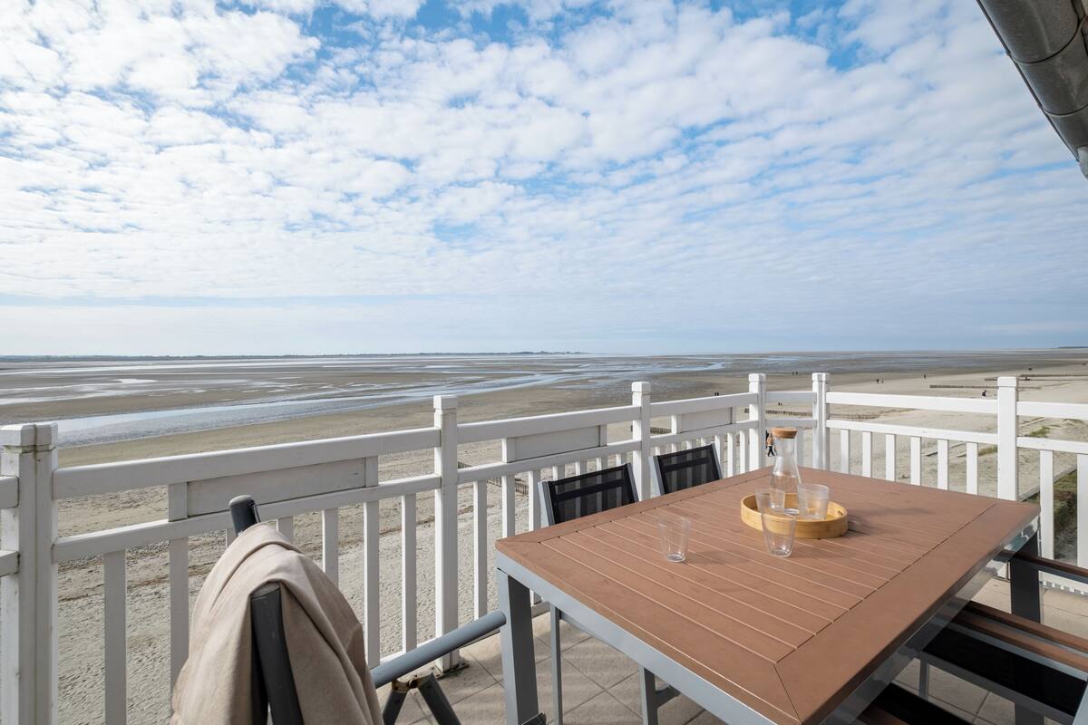 A wooden table with a bottle and glasses, a white railing, and a view of the beach.