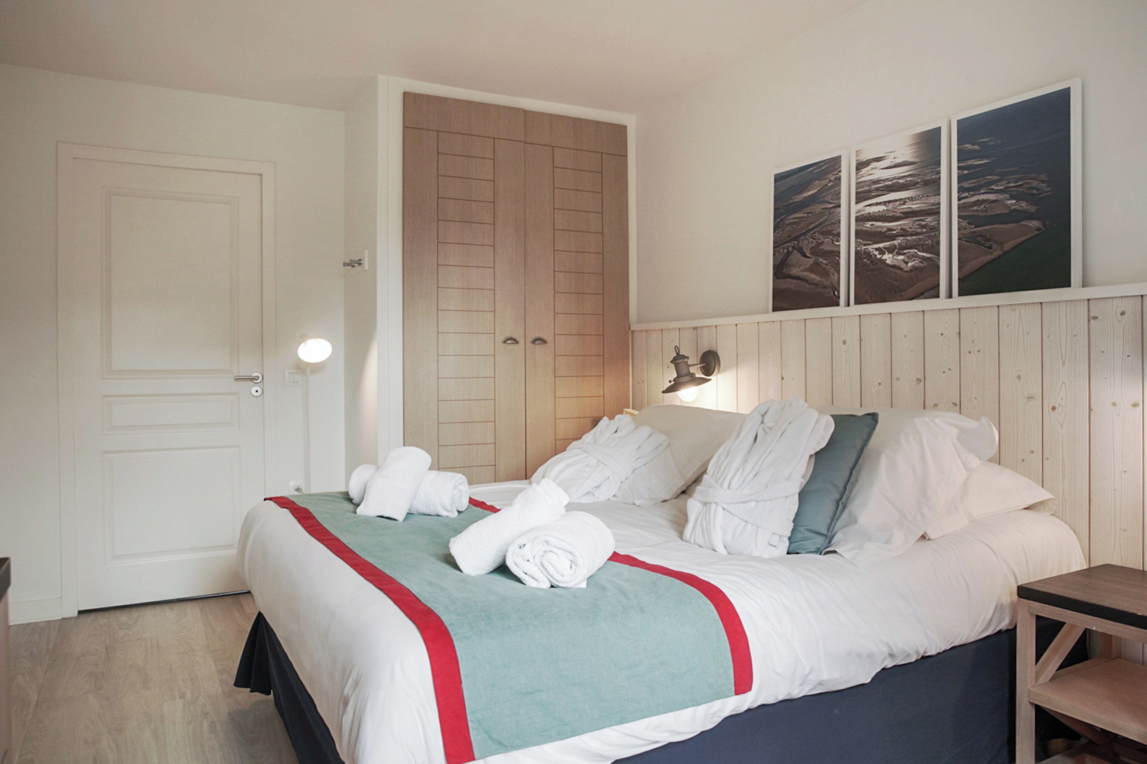 Un lit bien fait, avec du linge de lit blanc et un détail turquoise, une armoire en bois et une œuvre d’art accrochée au mur représentant un paysage.