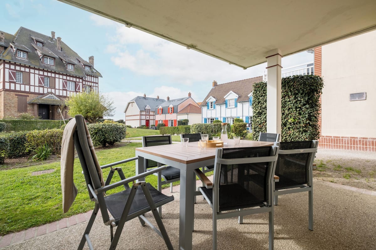 An outdoor patio with a table and chairs, surrounded by a garden and a row of colorful houses.
