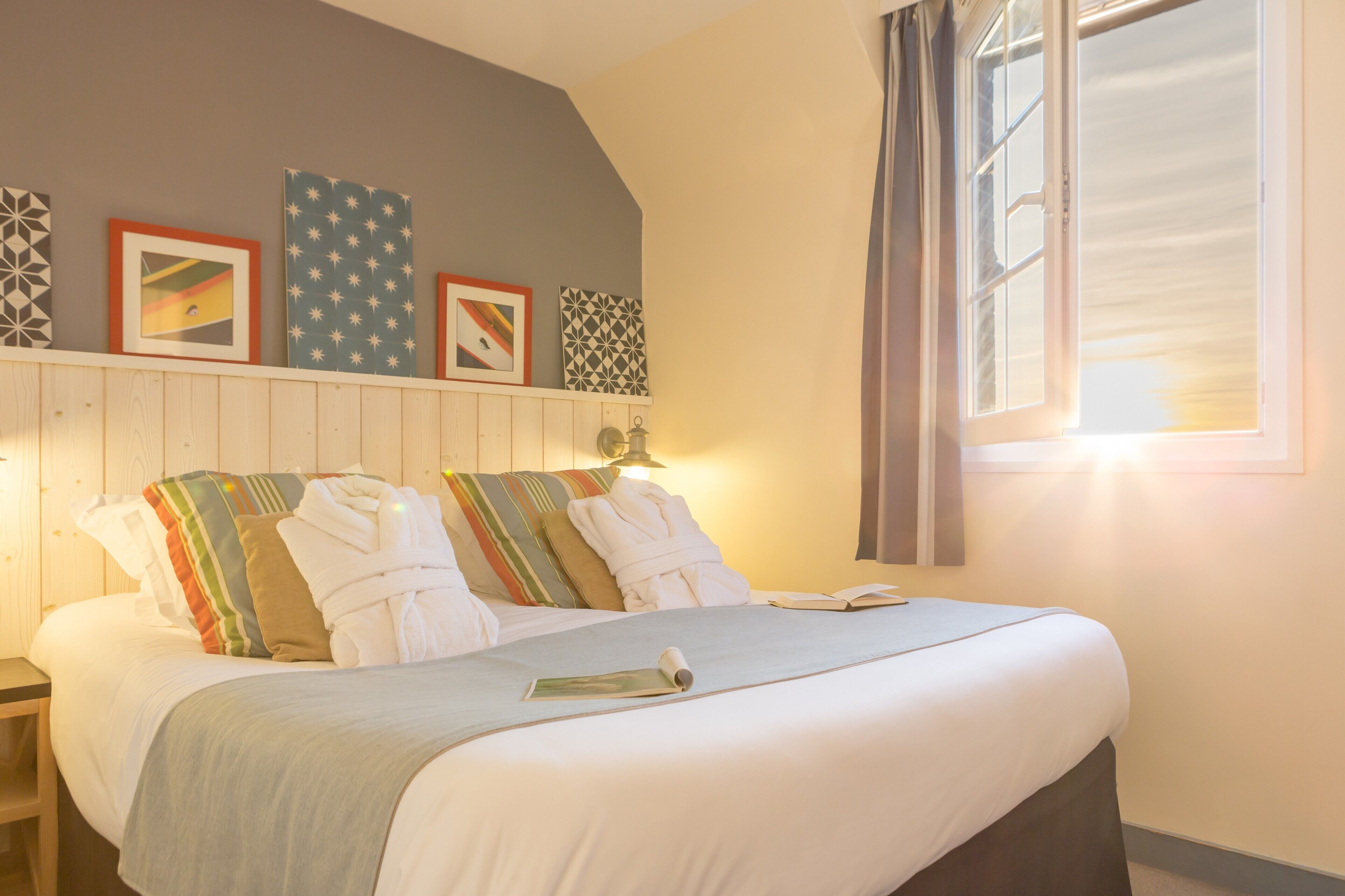 A neatly made bed with white linens and striped pillows, a bedside table with a lamp, and a window with curtains.