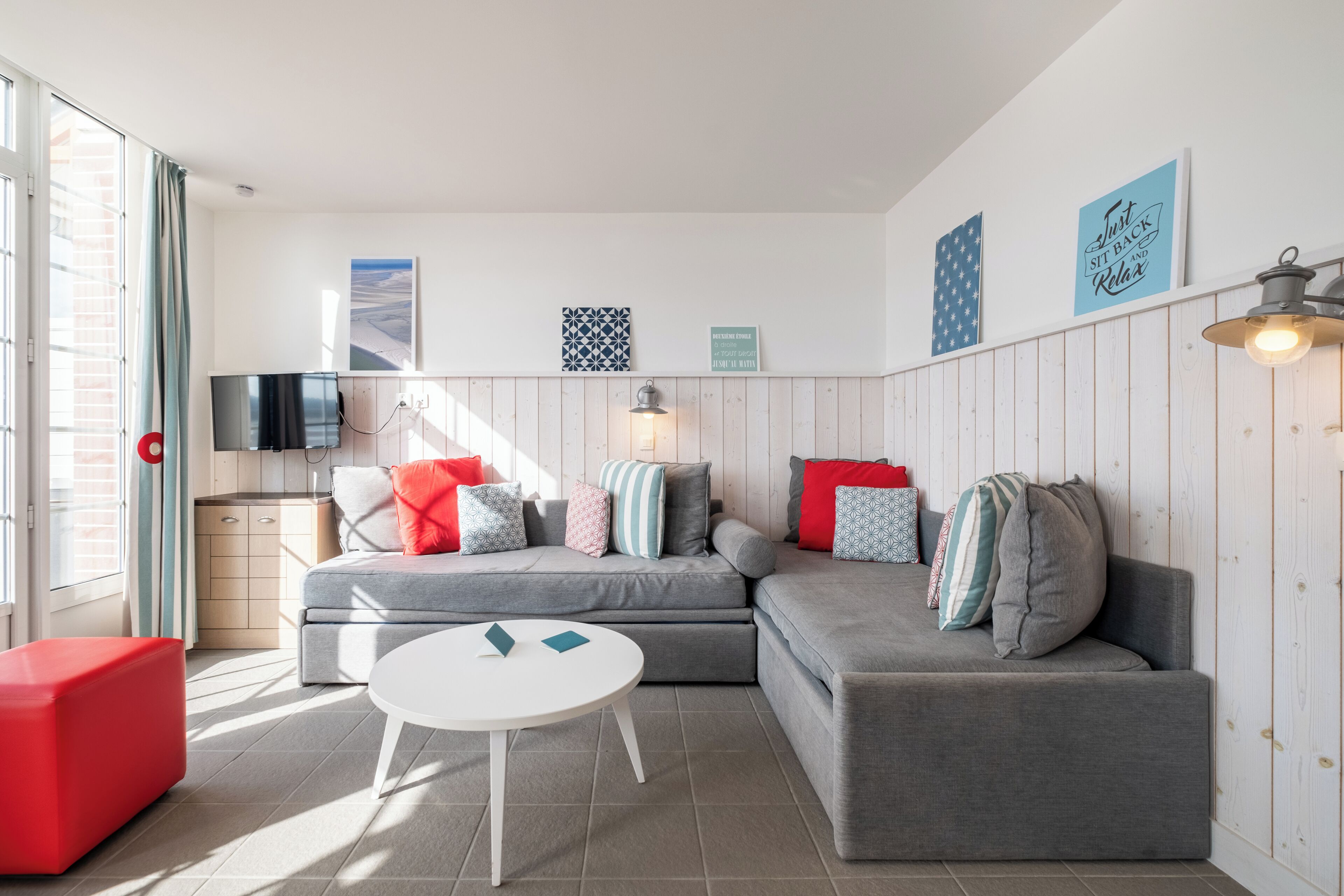 A modern living room with a grey sectional sofa, a white coffee table, and a red ottoman. There is a TV mounted on the wall, a window with blinds, and a wall-mounted lamp.