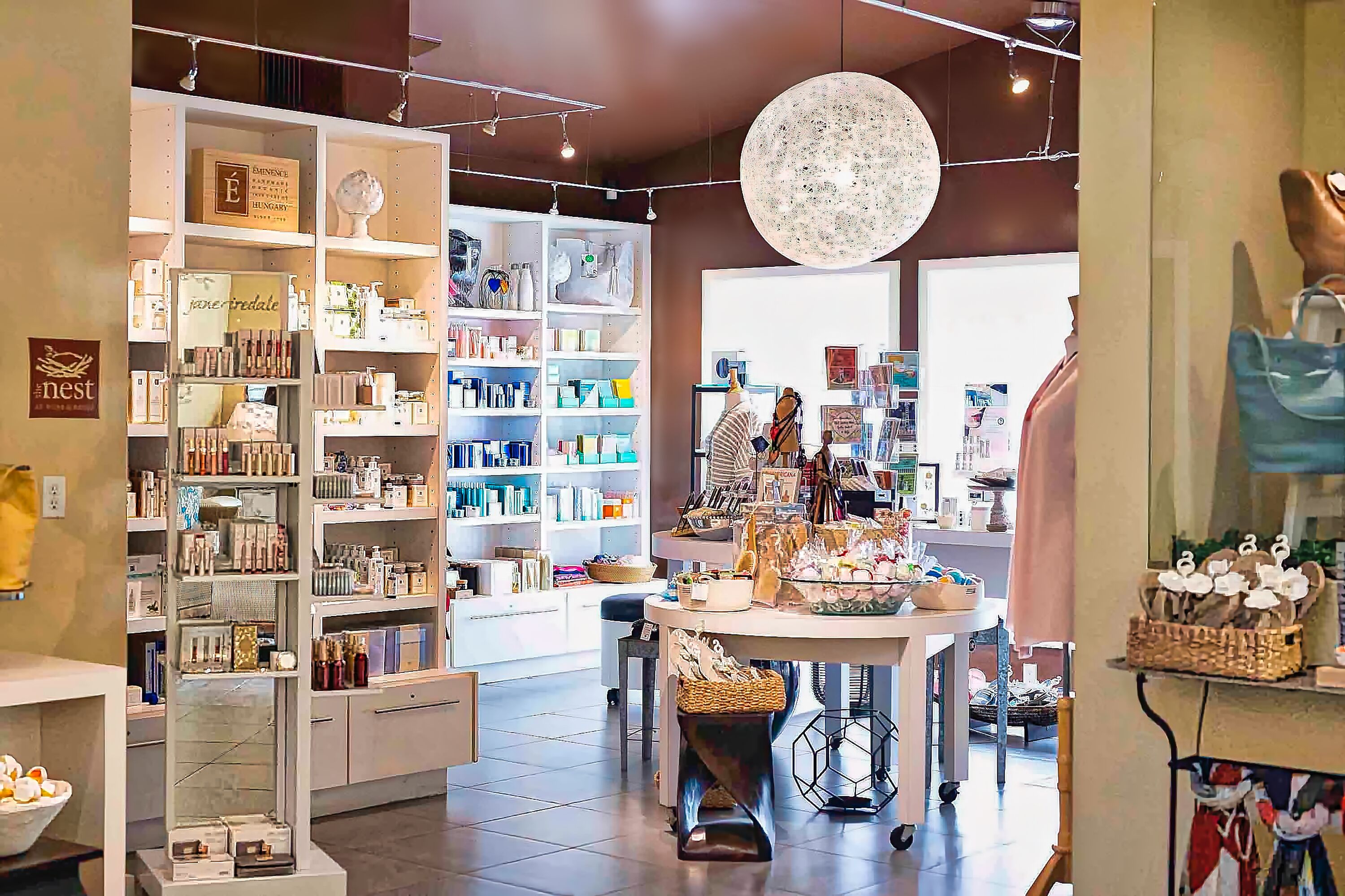 Una tienda boutique con estantes llenos de diversos productos, una mesa expositora central e iluminación colgante.