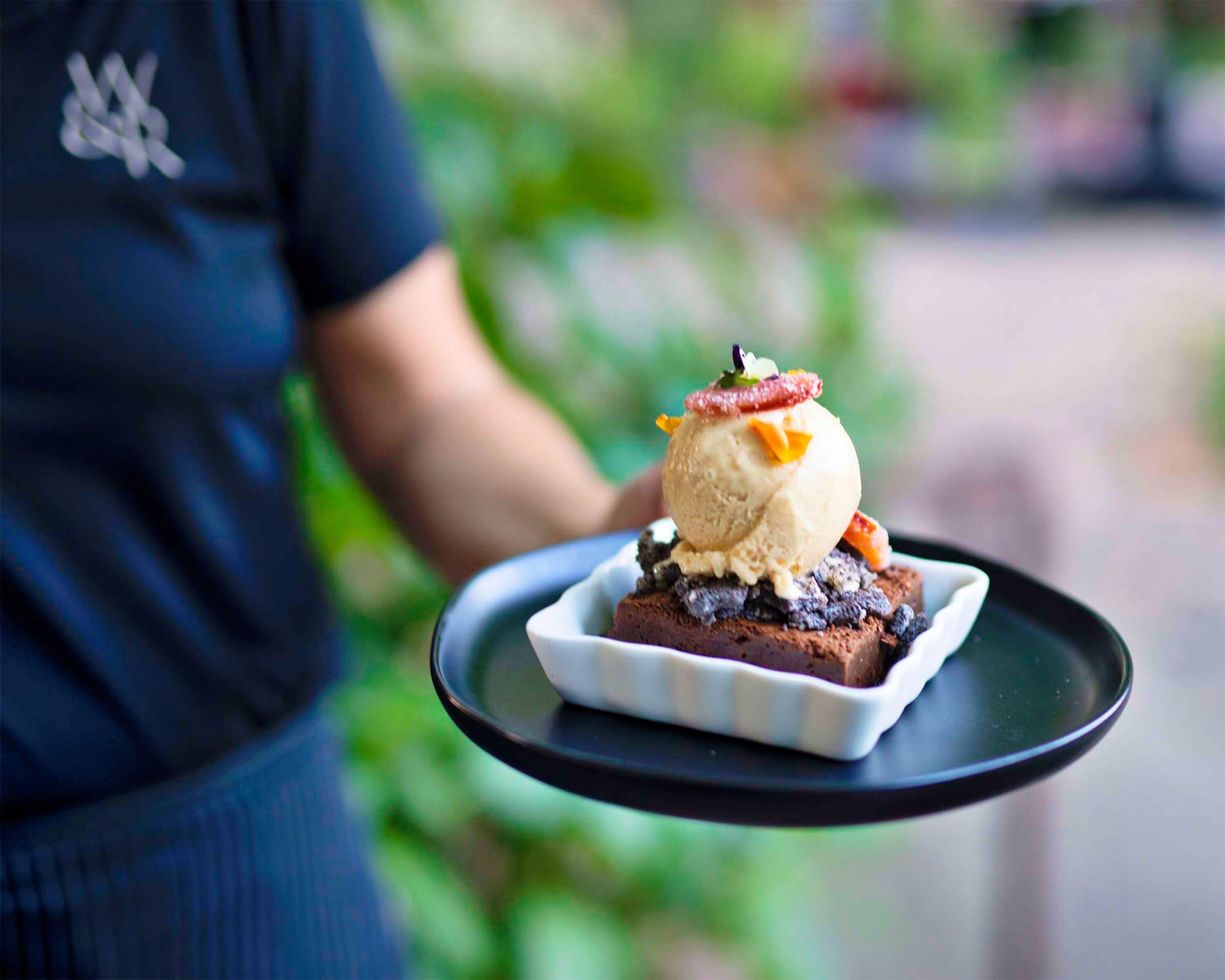 A dessert on a white square plate with a scoop of ice cream on top, garnished with fruit, served on a black tray.