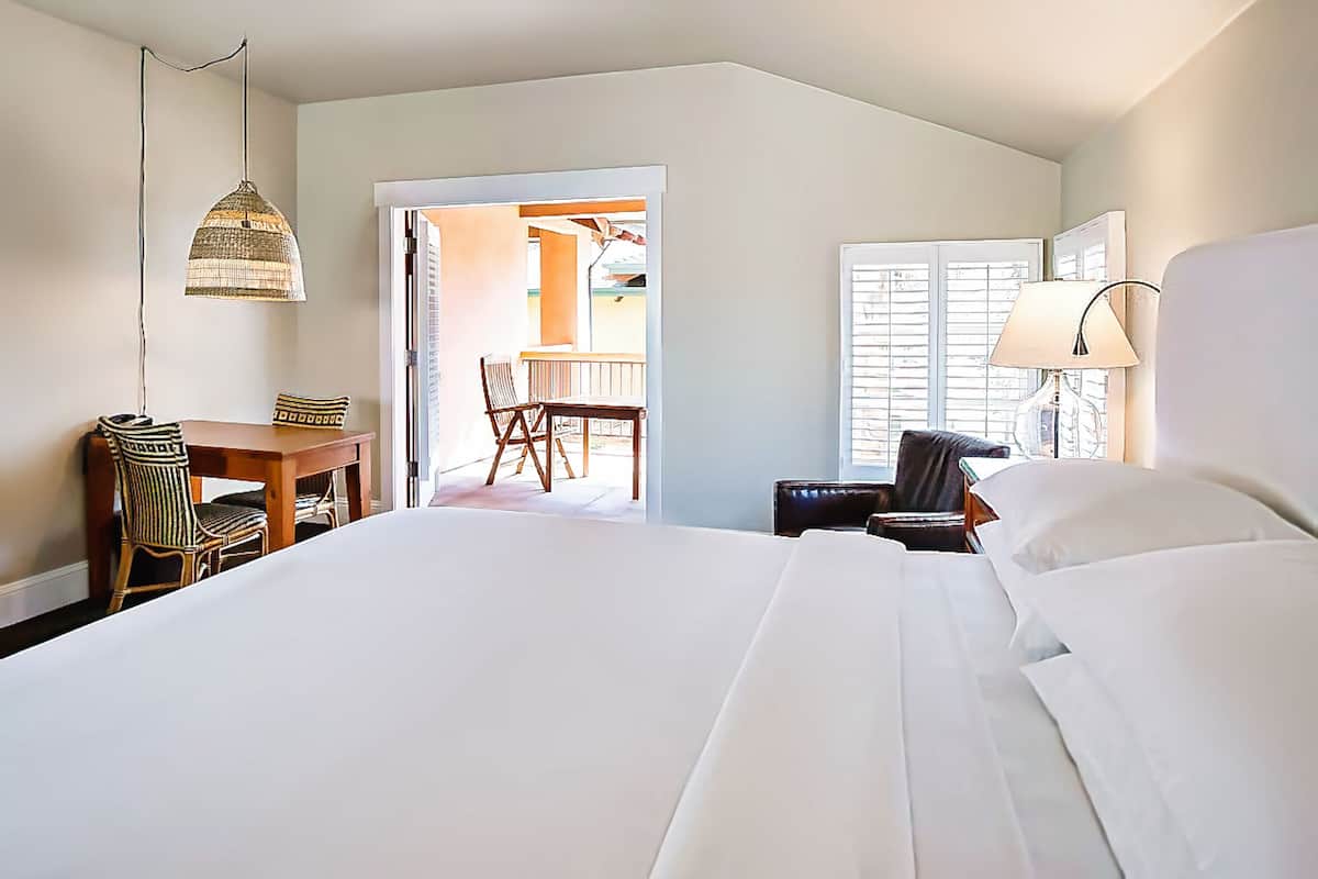 A hotel room with a large bed, a desk with a chair, a hanging lamp, and a view of an outdoor patio area.
