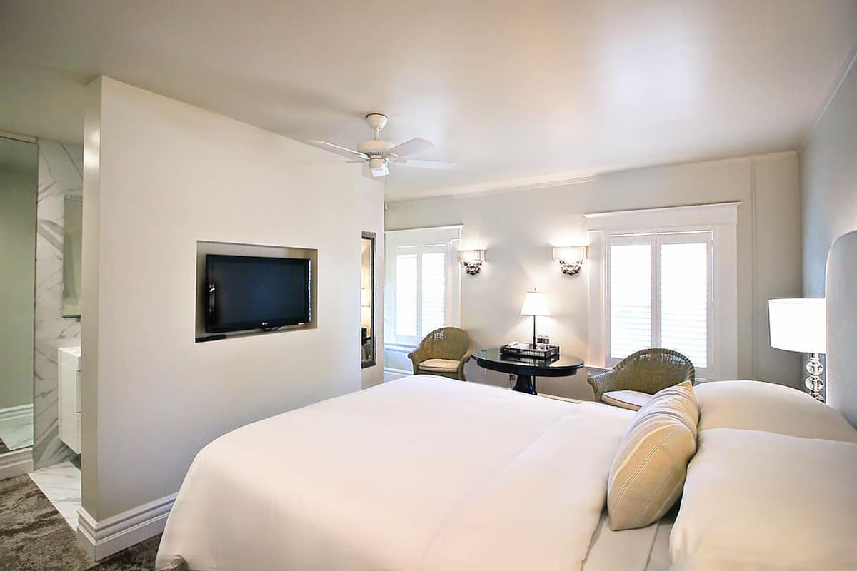 A hotel room with a large bed, a ceiling fan, a TV mounted on the wall, two chairs, a small table, and a lamp.