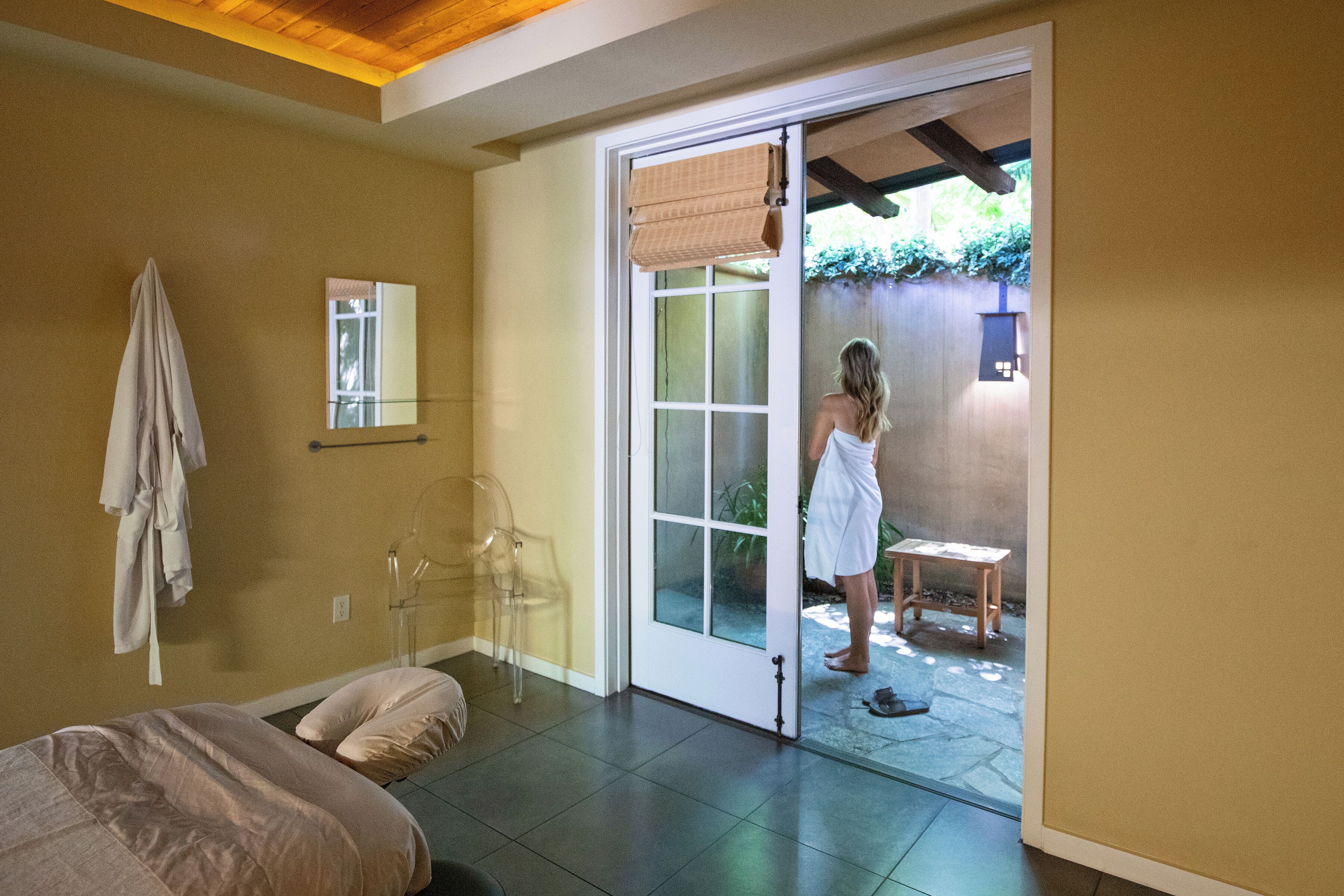 Una habitación con cama, baño con espejo y puerta corrediza de vidrio que da a un espacio exterior con mesa y sillas.