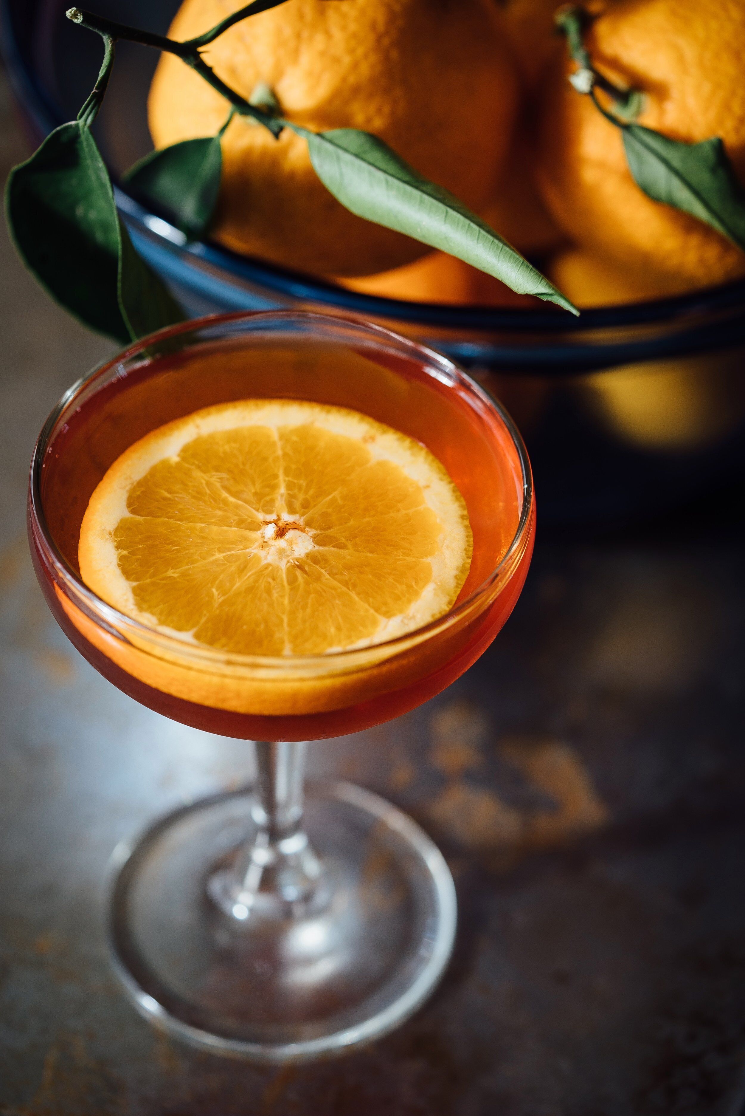 A cocktail glass with a slice of orange on the rim, placed on a table with a blurred background of oranges and leaves.