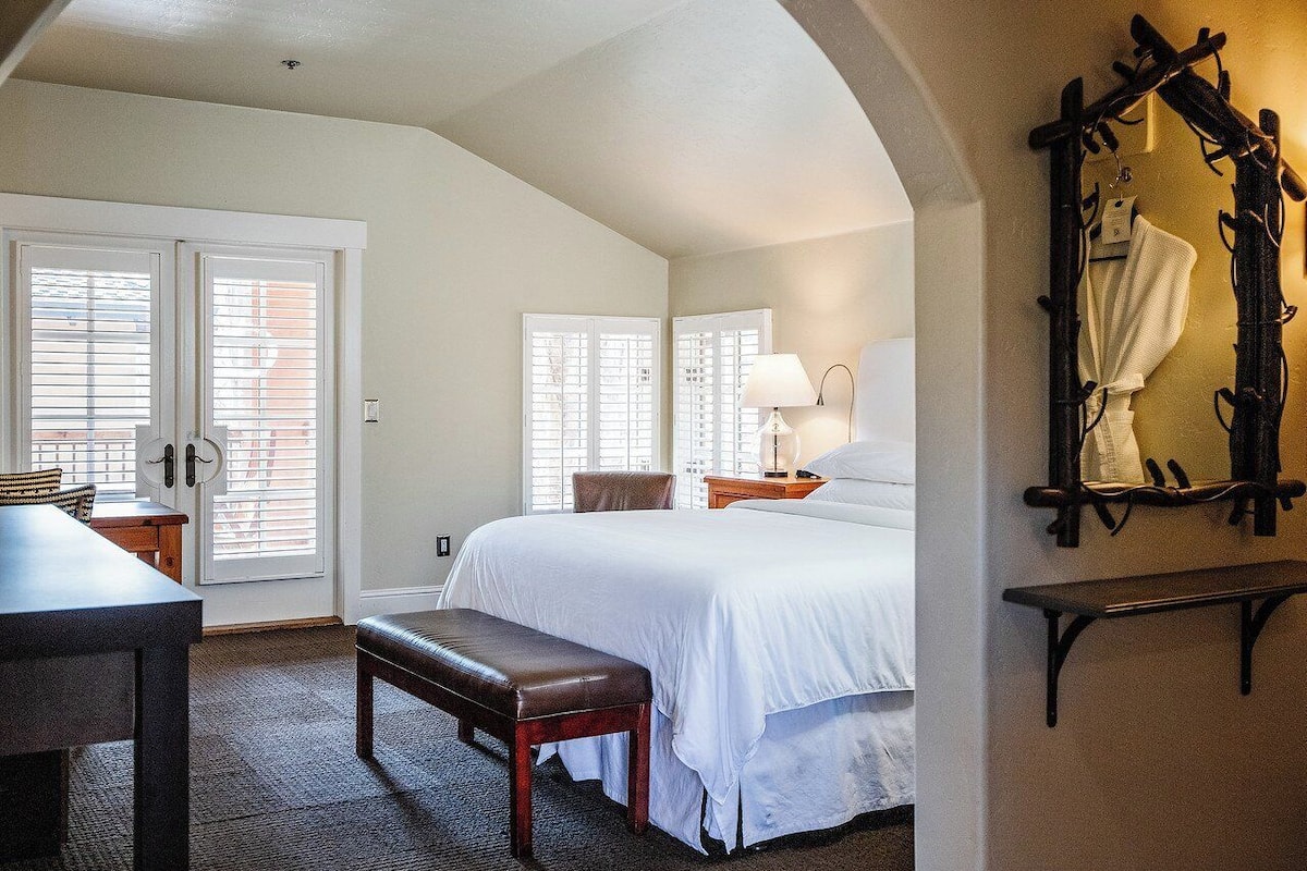 A bedroom with a large bed, two bedside tables with lamps, a bench, and a mirror with a towel hanging.