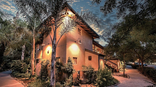 A two-story house with a well-lit exterior, surrounded by lush vegetation and a paved pathway.