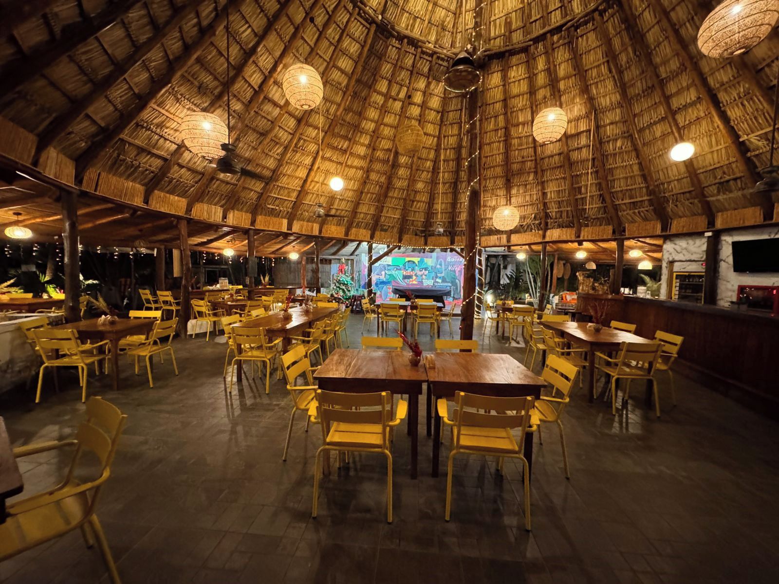 A restaurant with a thatched roof, wooden tables, and yellow chairs.