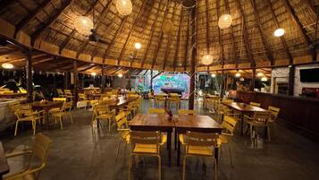 A restaurant with a thatched roof, wooden tables, and yellow chairs.
