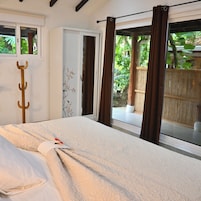 Quarto com cama, um cabideiro de madeira, janela com cortinas e vista para o verde lá fora.