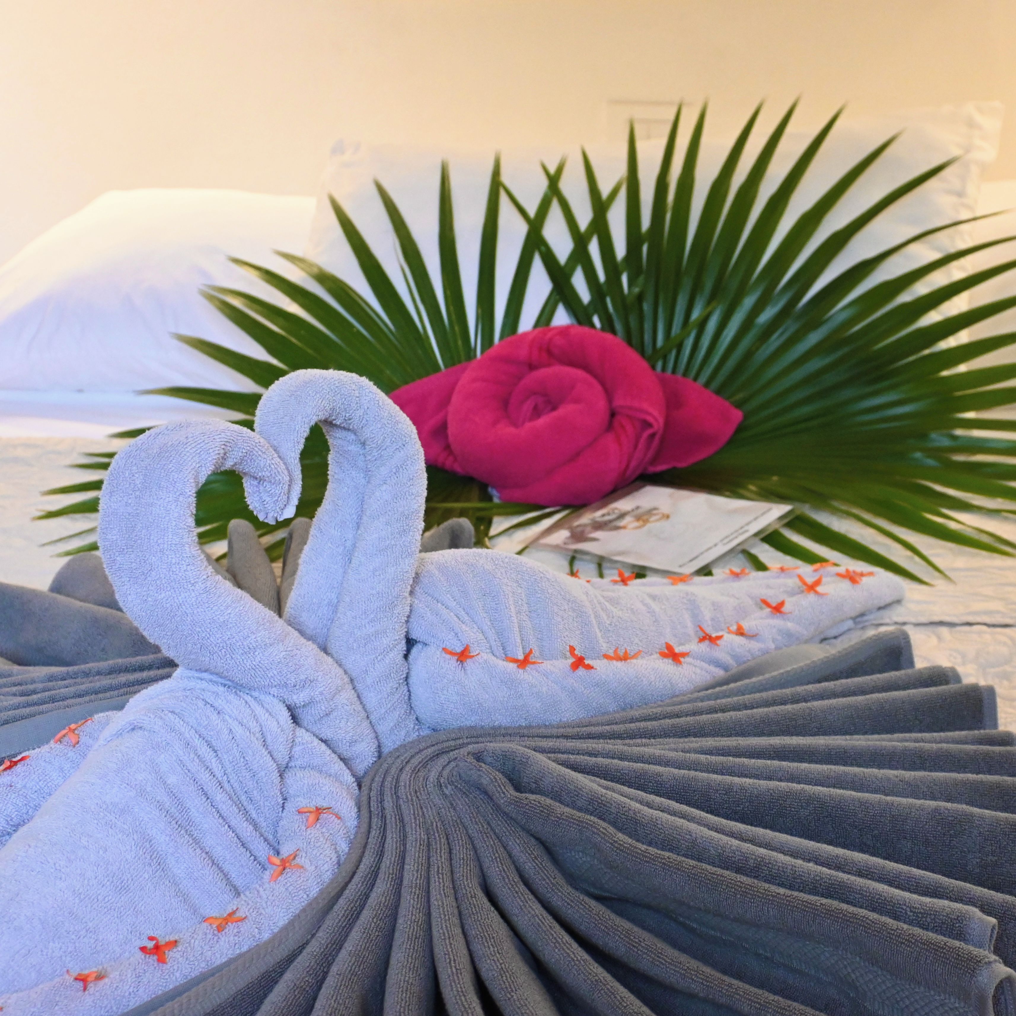 A neatly arranged set of towels with a heart-shaped towel arrangement, a pink rose, and a green palm leaf in the background.
