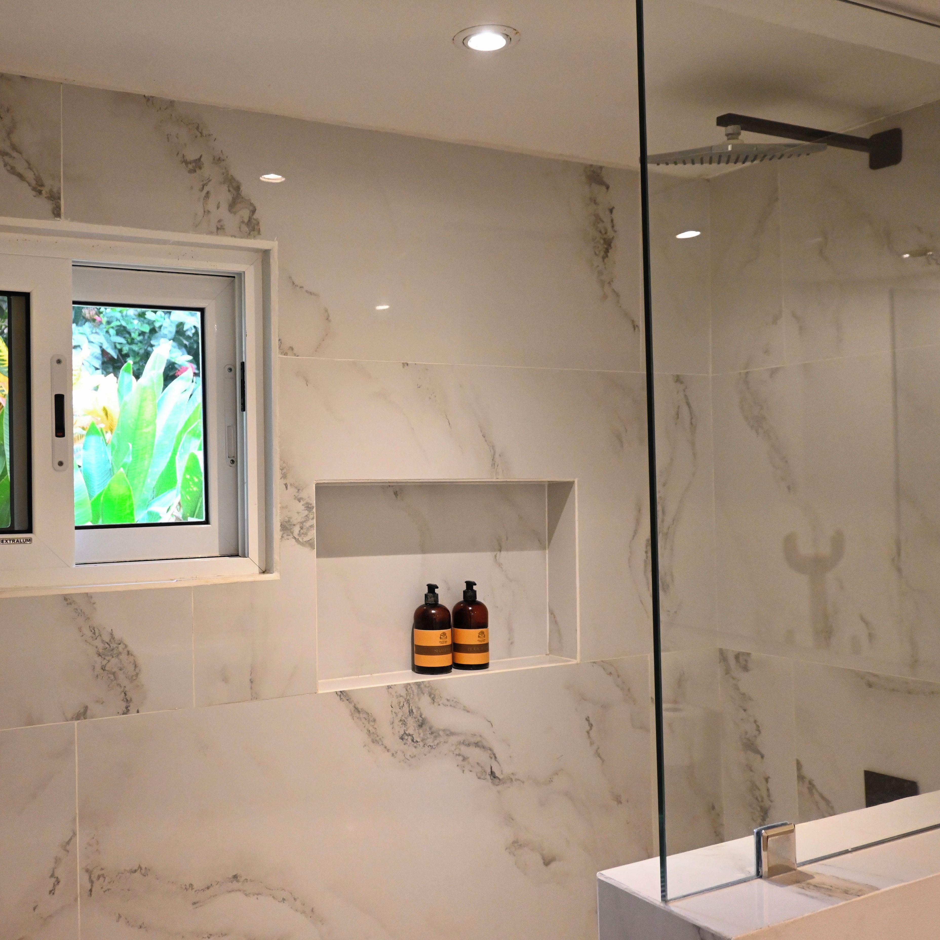 Un baño con pared de mármol, una ventana con vistas a la vegetación y dos botellas sobre una estantería.