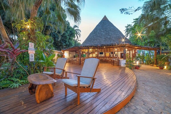 Una terraza de madera con sillas y una mesa, rodeada de vegetación tropical y una estructura con techo de paja.