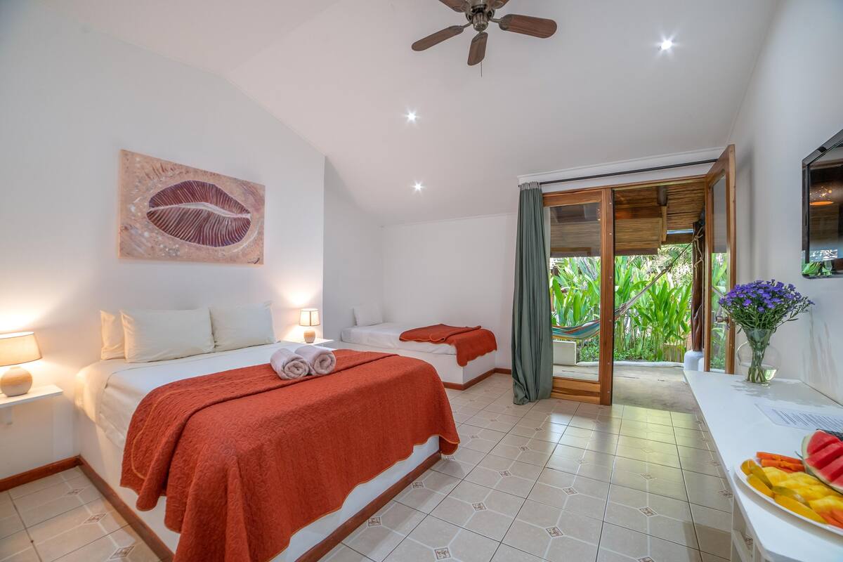 A hotel room with two beds, a ceiling fan, a large window with curtains, a small table with a vase of flowers, and a fruit bowl on the side.