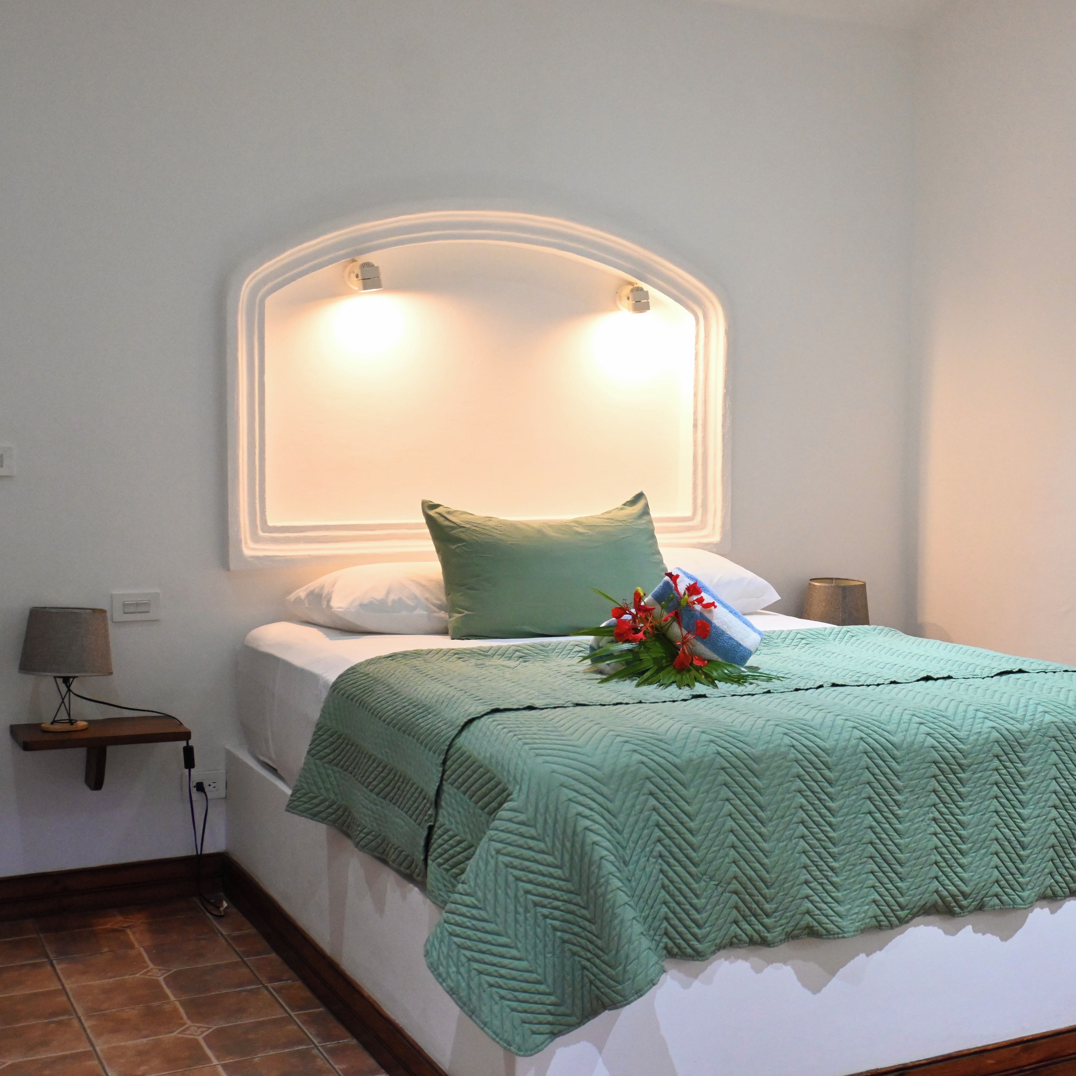 Ein ordentlich gemachtes Bett mit grüner Decke und Kissen, einer Nachttischlampe und einer Wandleuchte.