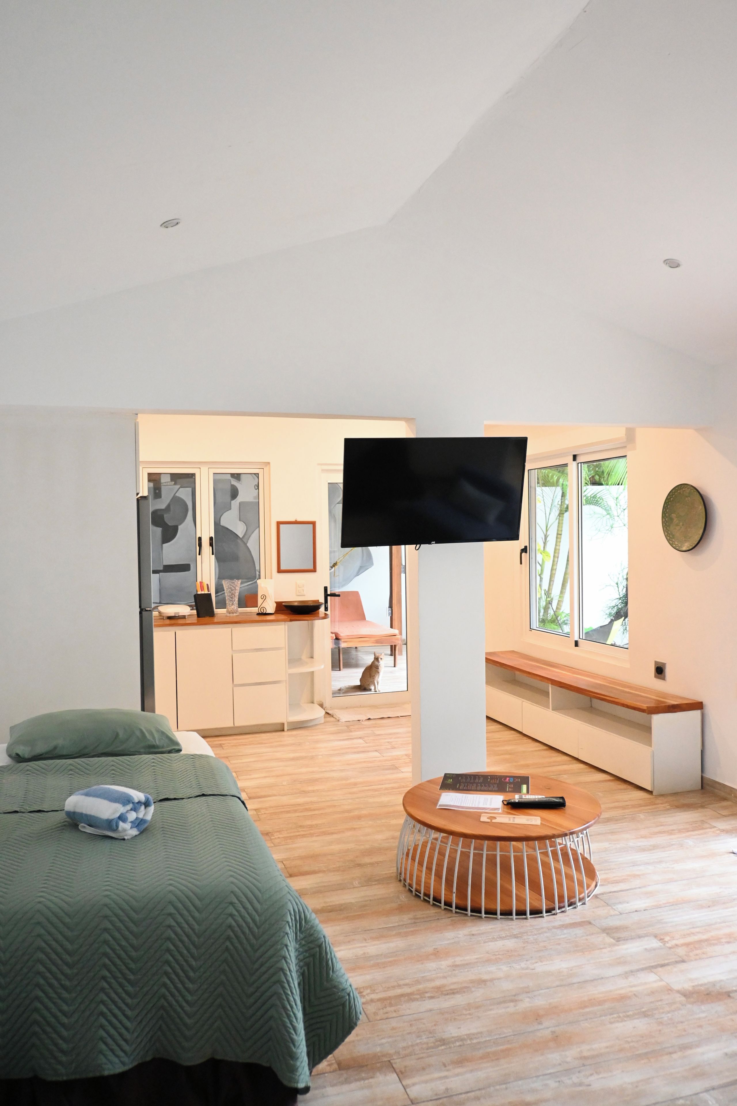 Un dormitorio moderno con una cama, una mesa redonda de madera, un televisor y vistas a una cocina y zona de comedor.