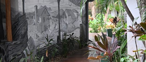 A pathway leading through a lush garden with a mural on the wall depicting a rural scene.
