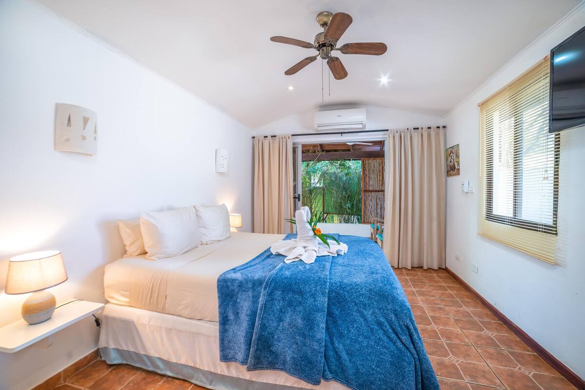 A bedroom with a bed, a ceiling fan, a window with blinds, and a wall-mounted air conditioner.