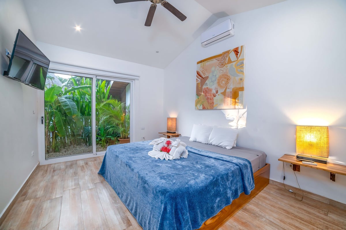 A bedroom with a bed, a TV, a ceiling fan, a window with a view of greenery, and a wall-mounted air conditioner.